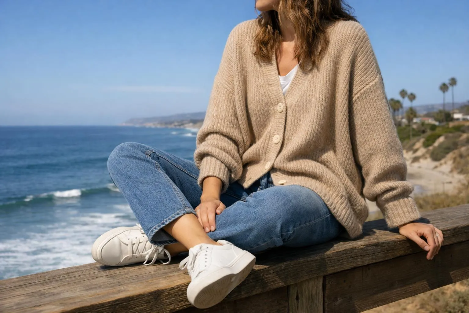 Femme assise sur une jetée, admirant l'océan, vêtue d'un cardigan beige.