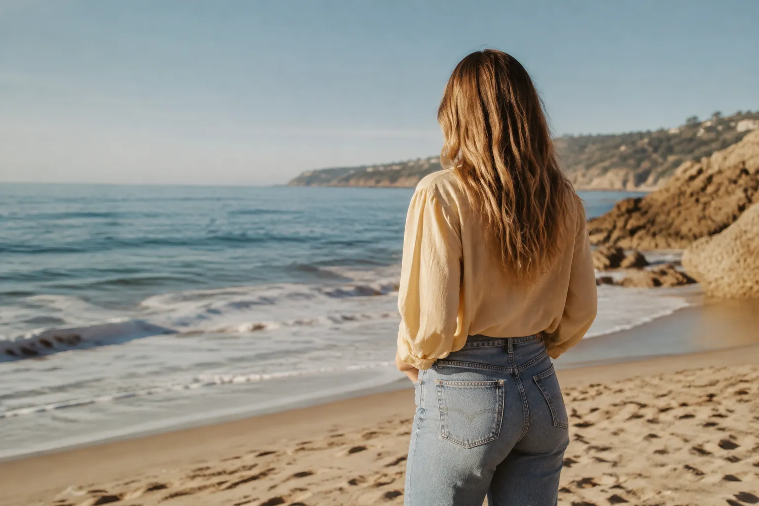 Femme aux cheveux longs, debout sur une plage rocheuse, contemplant l'océan.