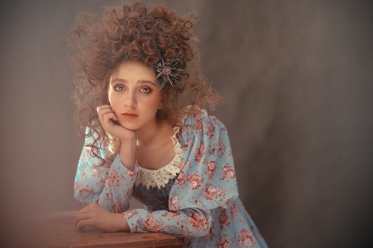Femme aux cheveux bouclés, robe fleurie, fleur dans les cheveux, regard pensif.