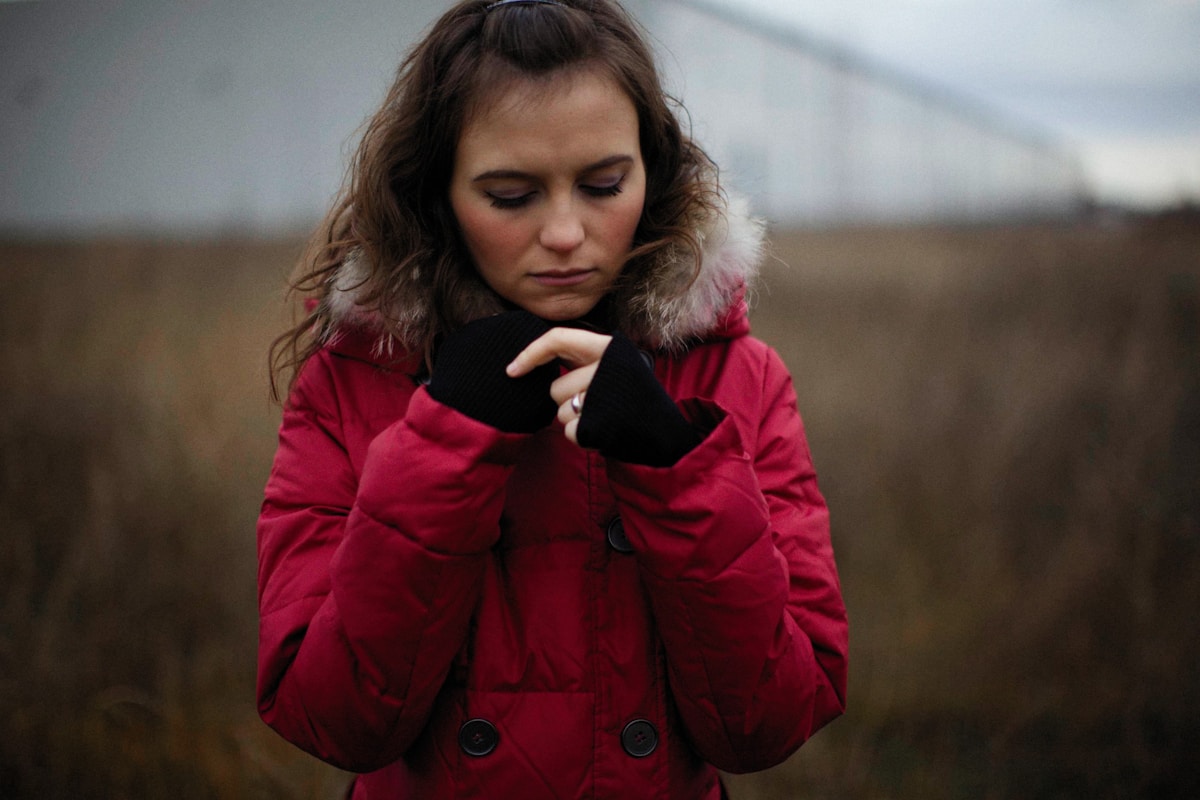 Femme en manteau rouge avec capuche, l'air pensive, dans un paysage flou.