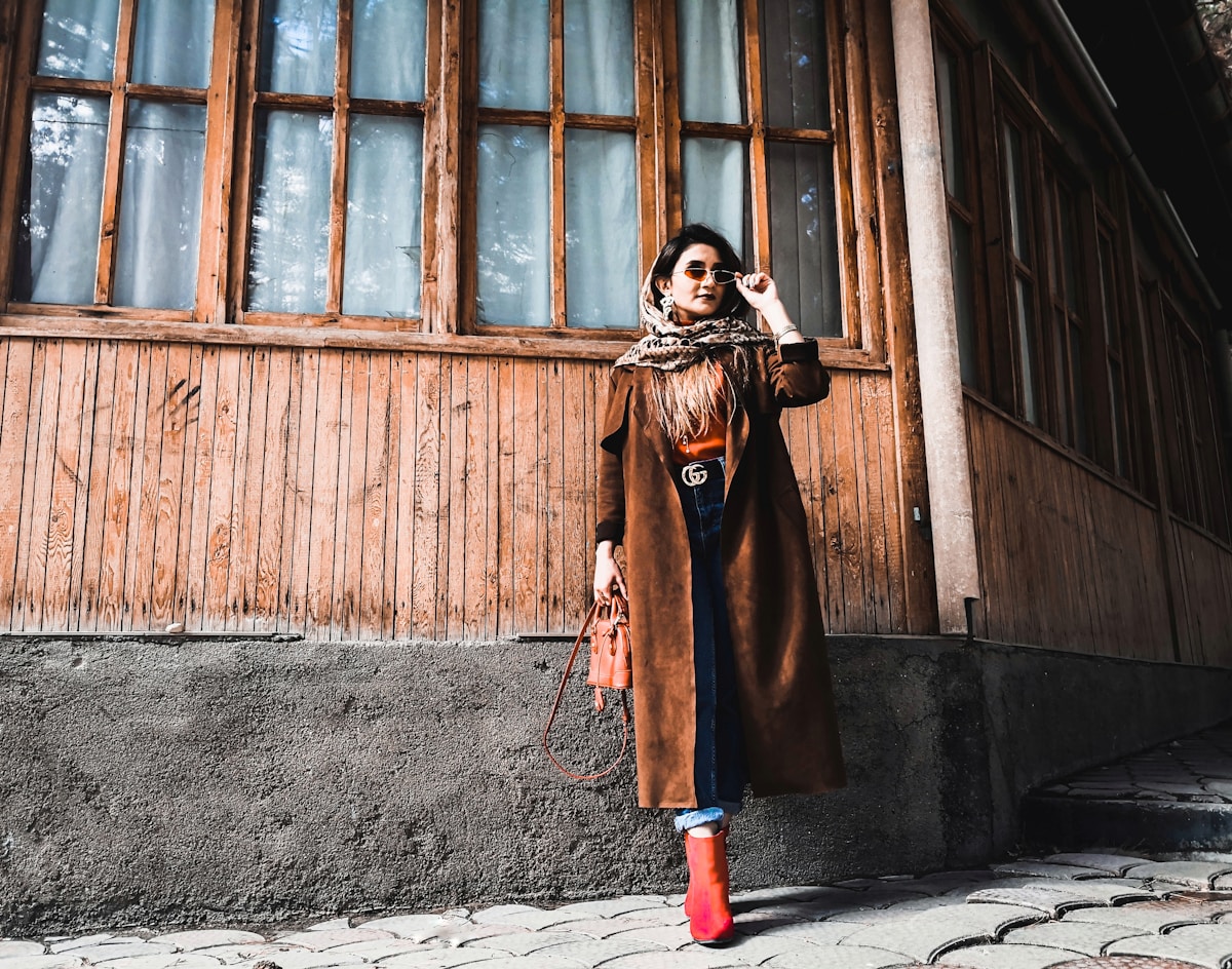 Femme élégante en manteau marron, écharpe et lunettes de soleil, debout devant une façade en bois.