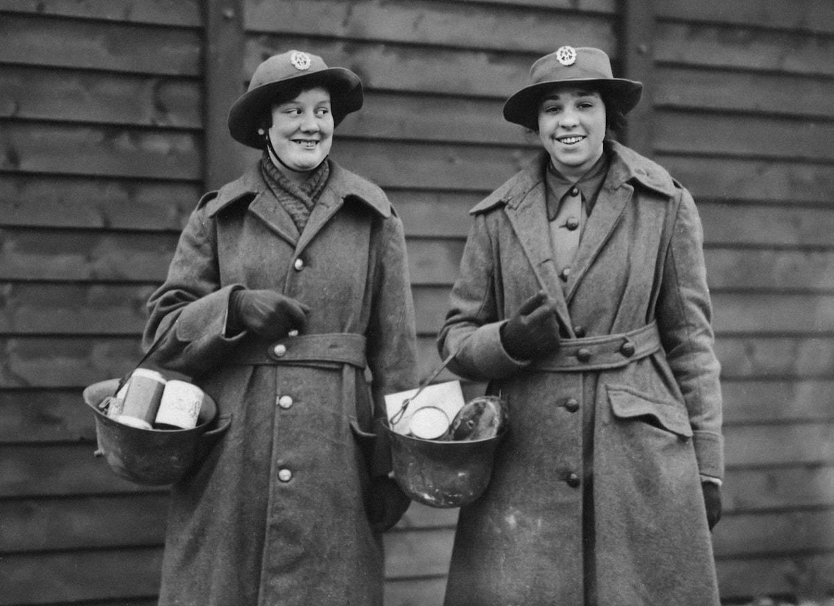Deux femmes en manteaux et chapeaux souriant devant un bâtiment en bois.
