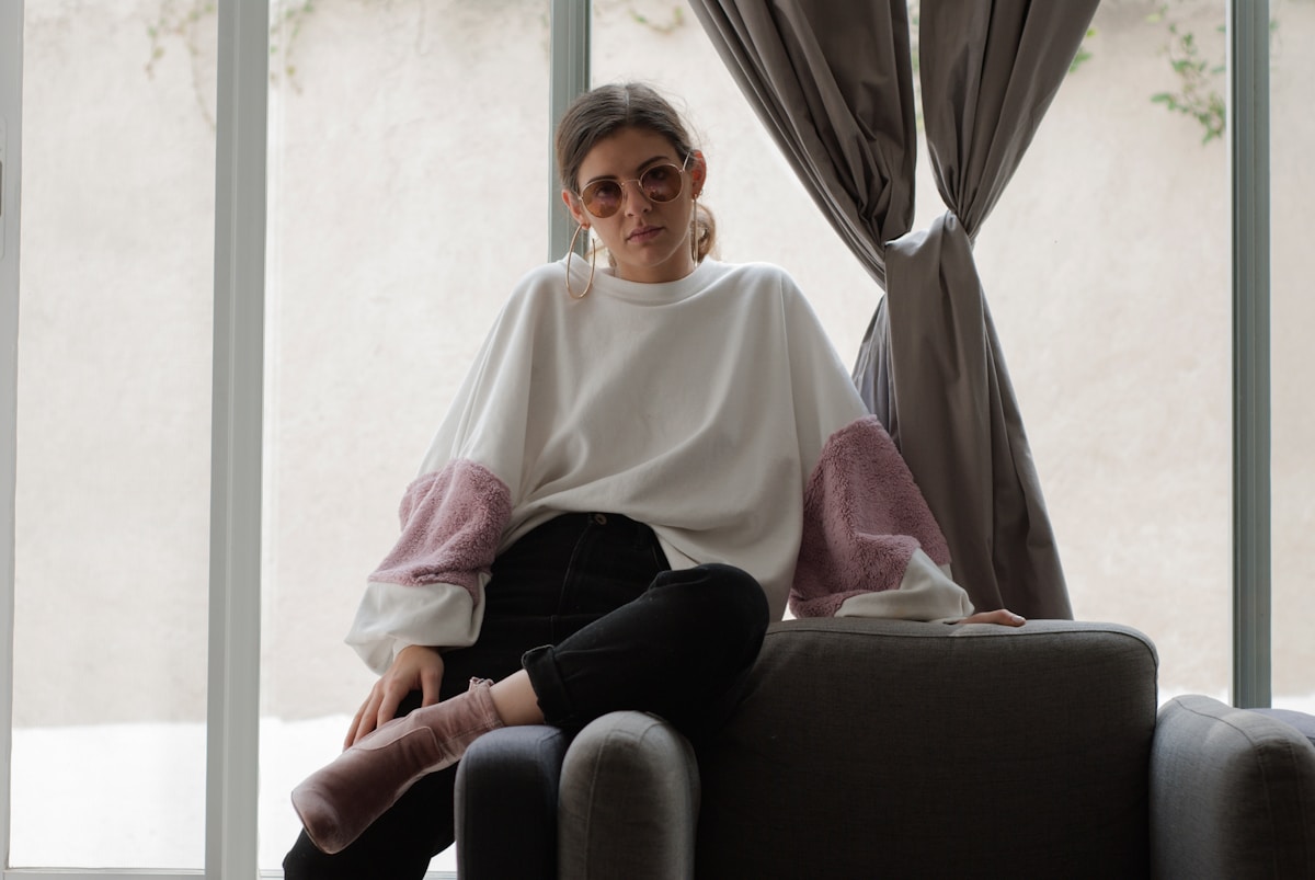 Femme assise sur un fauteuil, vêtue d'un pull blanc et de lunettes rondes.