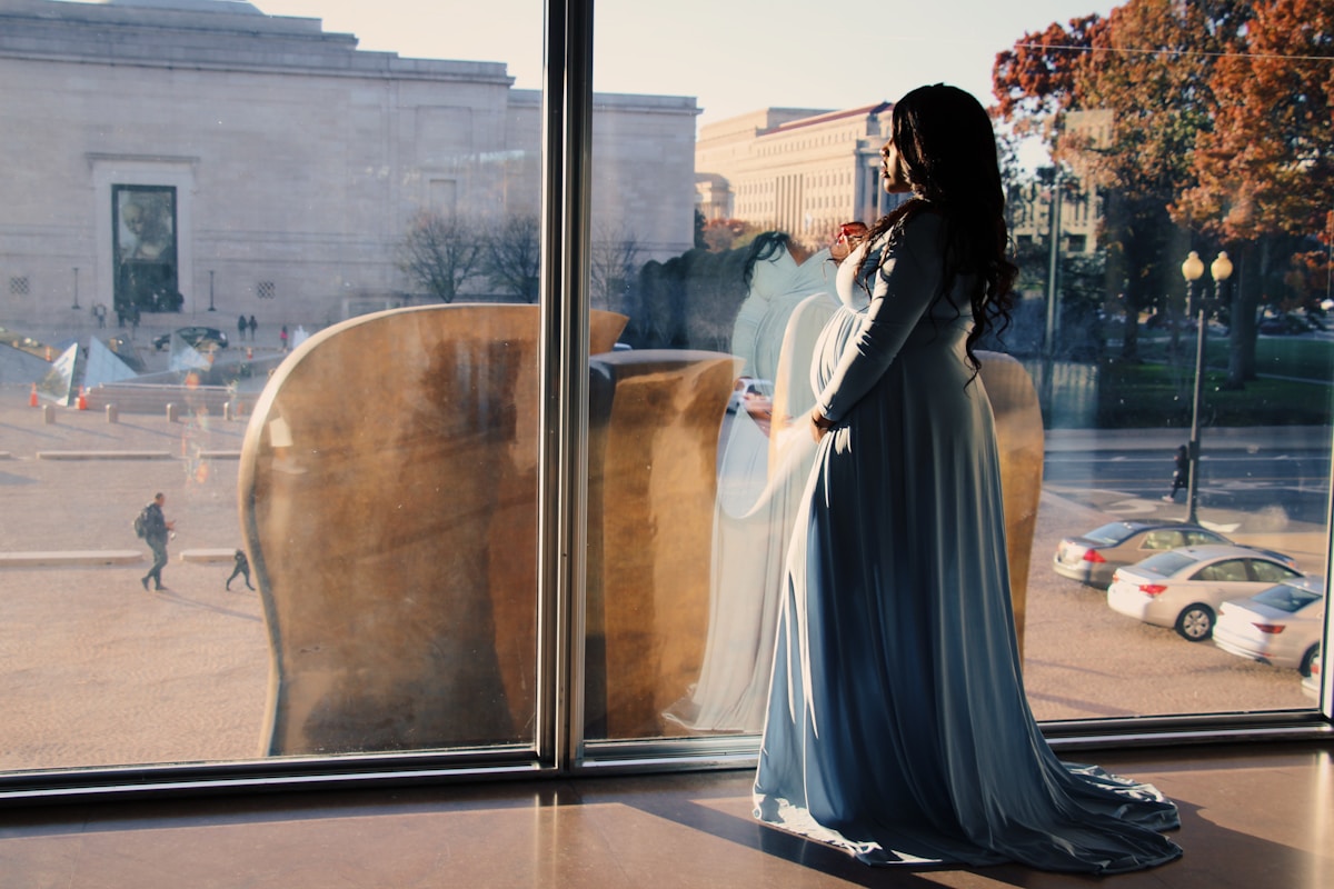 Femme enceinte en robe de soirée bleue, debout devant une fenêtre avec vue sur la ville.