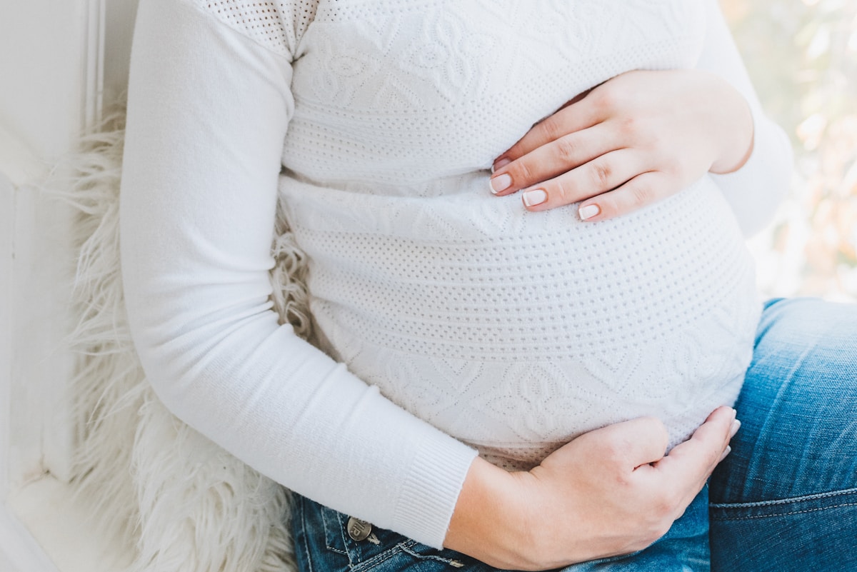 Femme enceinte vêtue d'un pull blanc, assise sur un sol moelleux.