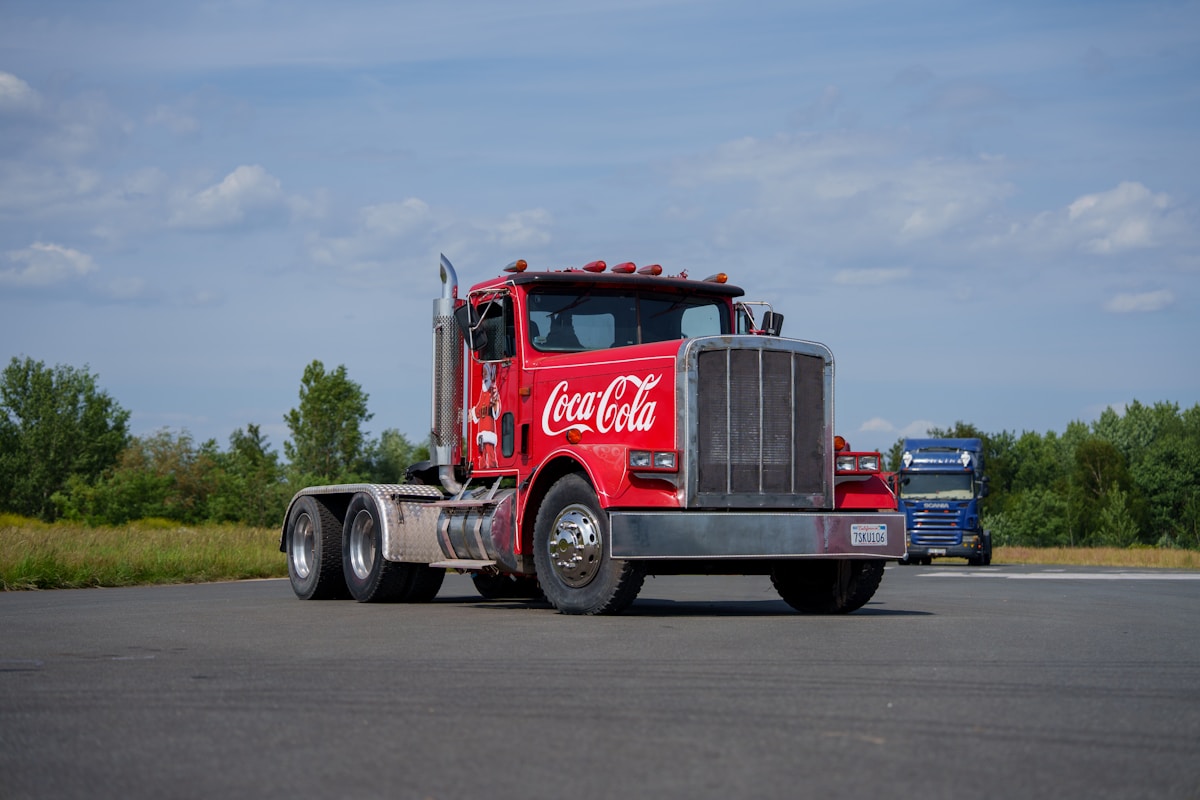 Camion rouge Coca-Cola sur une route goudronnée, entouré de verdure.