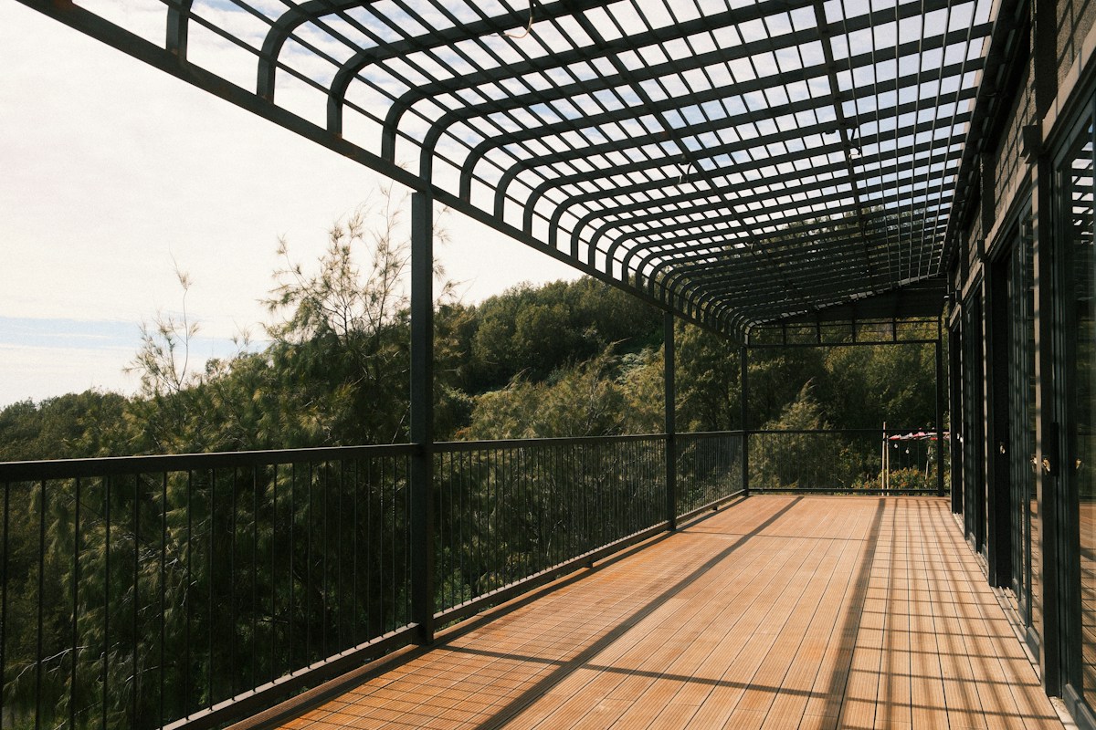 Terrasse en bois couverte d’une pergola métallique, entourée de végétation.