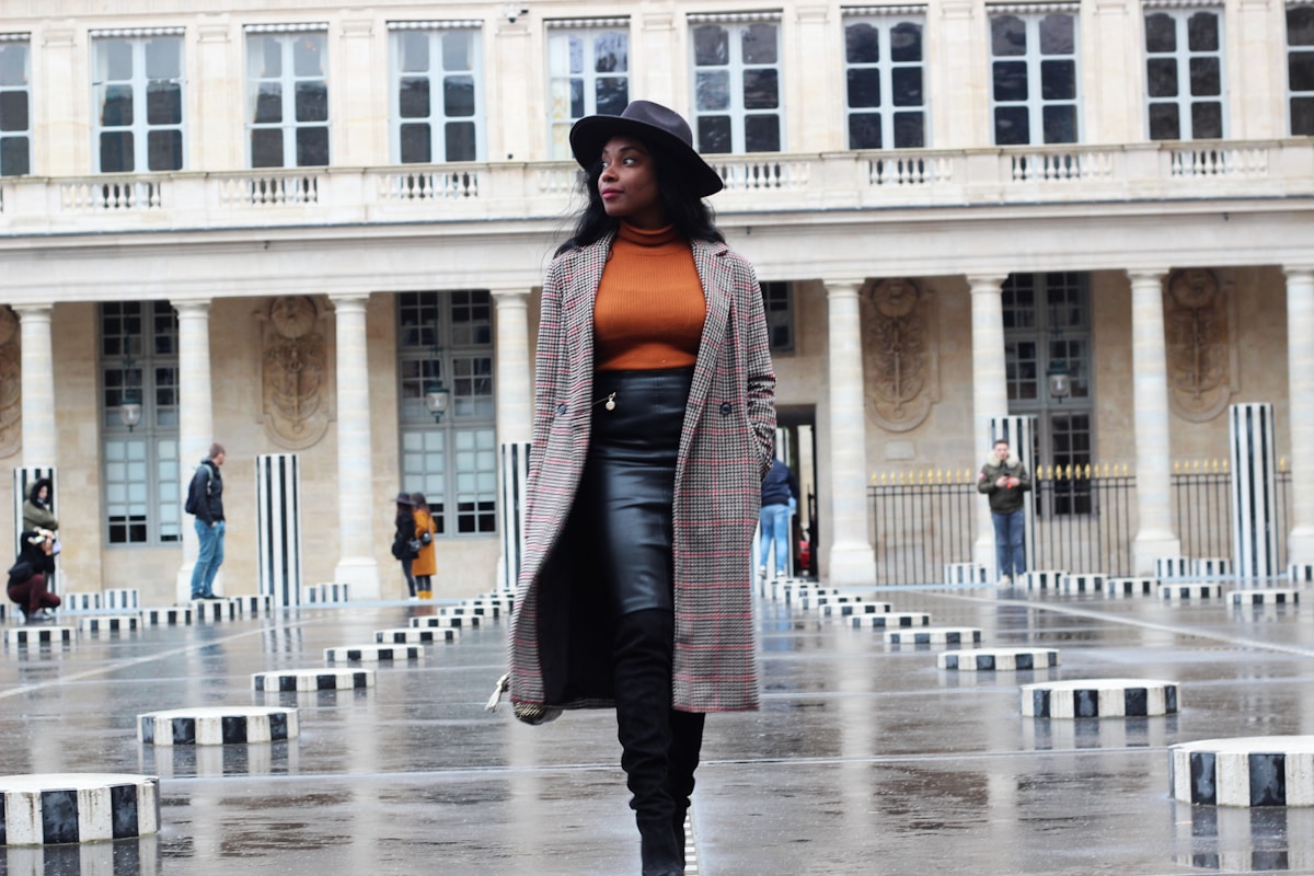 Femme élégante en manteau à carreaux, chapeau et bottes noires, debout dans une place publique.