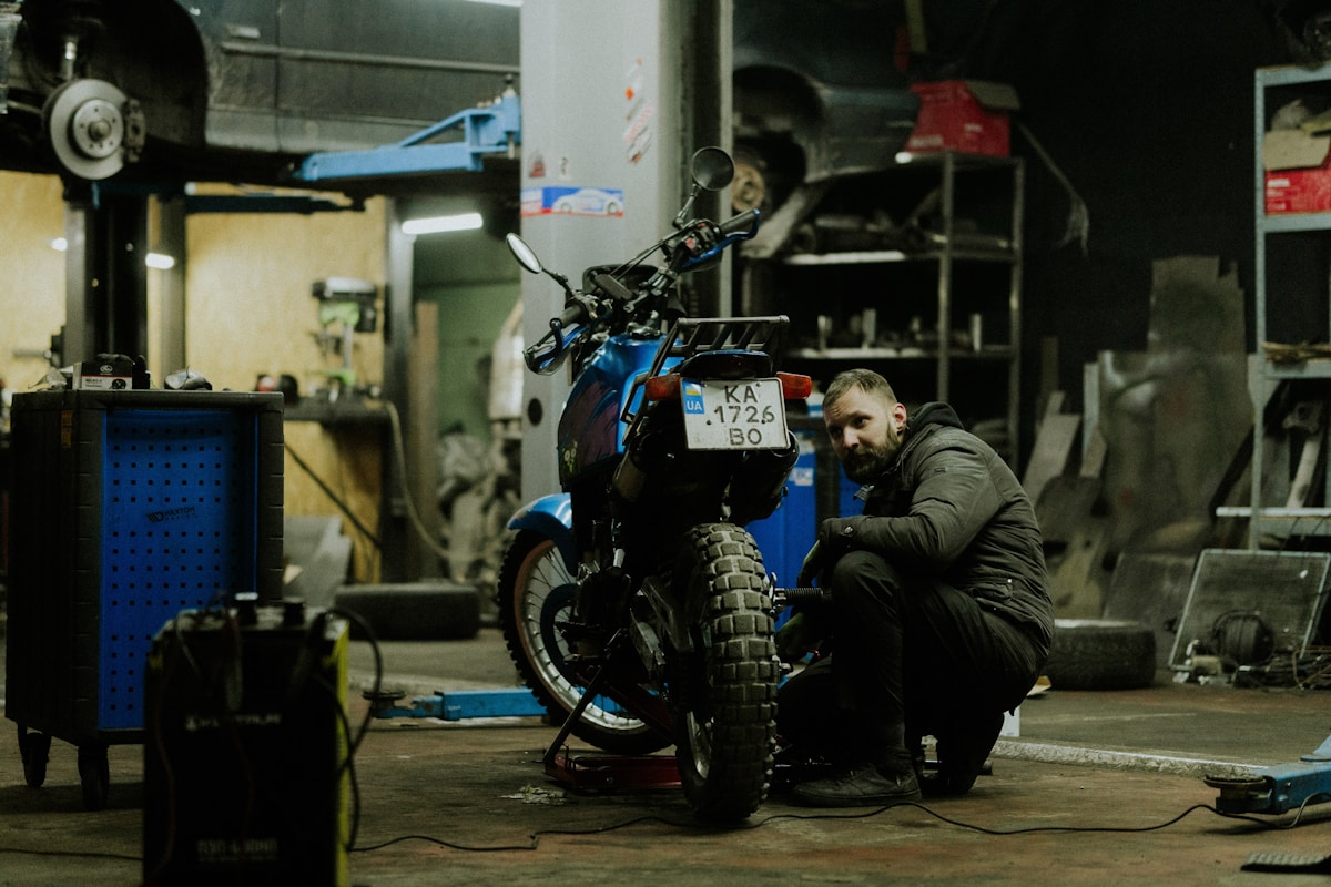 Un homme barbu répare une moto bleue dans un atelier sombre.