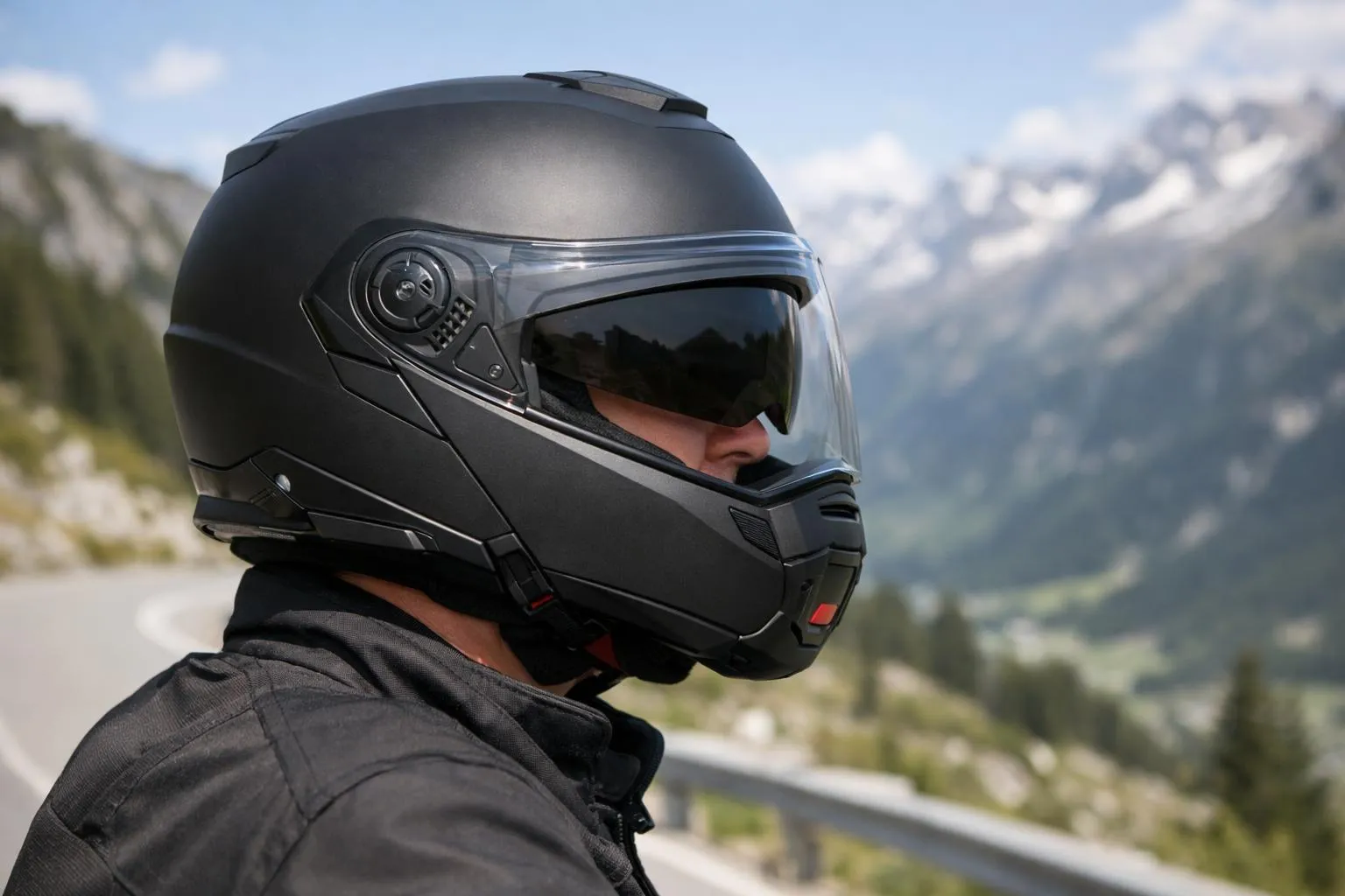 Motard portant un casque modulable Nolan N100-5 noir mat lors d'un arrêt sur une route de montagne suisse, vue de profil rapprochée montrant les détails de la mentonnière relevée et la visière pare-soleil, arrière-plan de paysage alpin flou, éclairage naturel