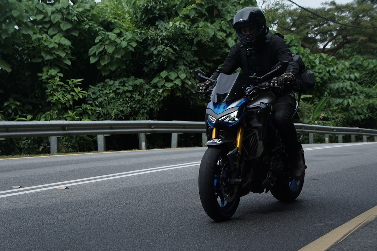 Motocycliste en équipement noir roulant sur une route bordée de végétation.