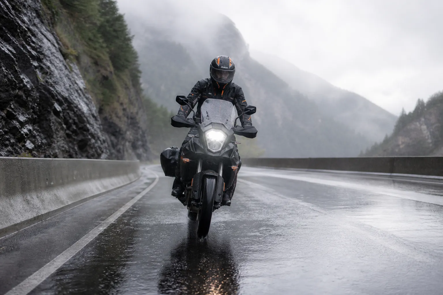 Motocycliste traversant une route de montagne sous la pluie.