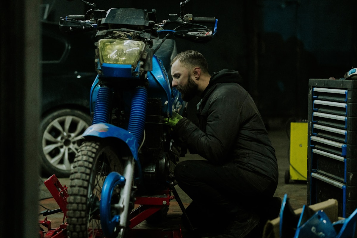 Homme barbu réparant une moto bleue dans un atelier.