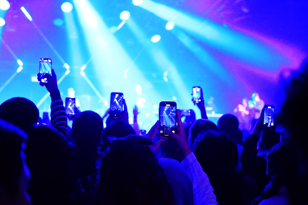 A crowd of people at a small, intimate concert. They are all looking towards the stage, and many are holding up their glowing smartphones, possibly recording or interacting online.