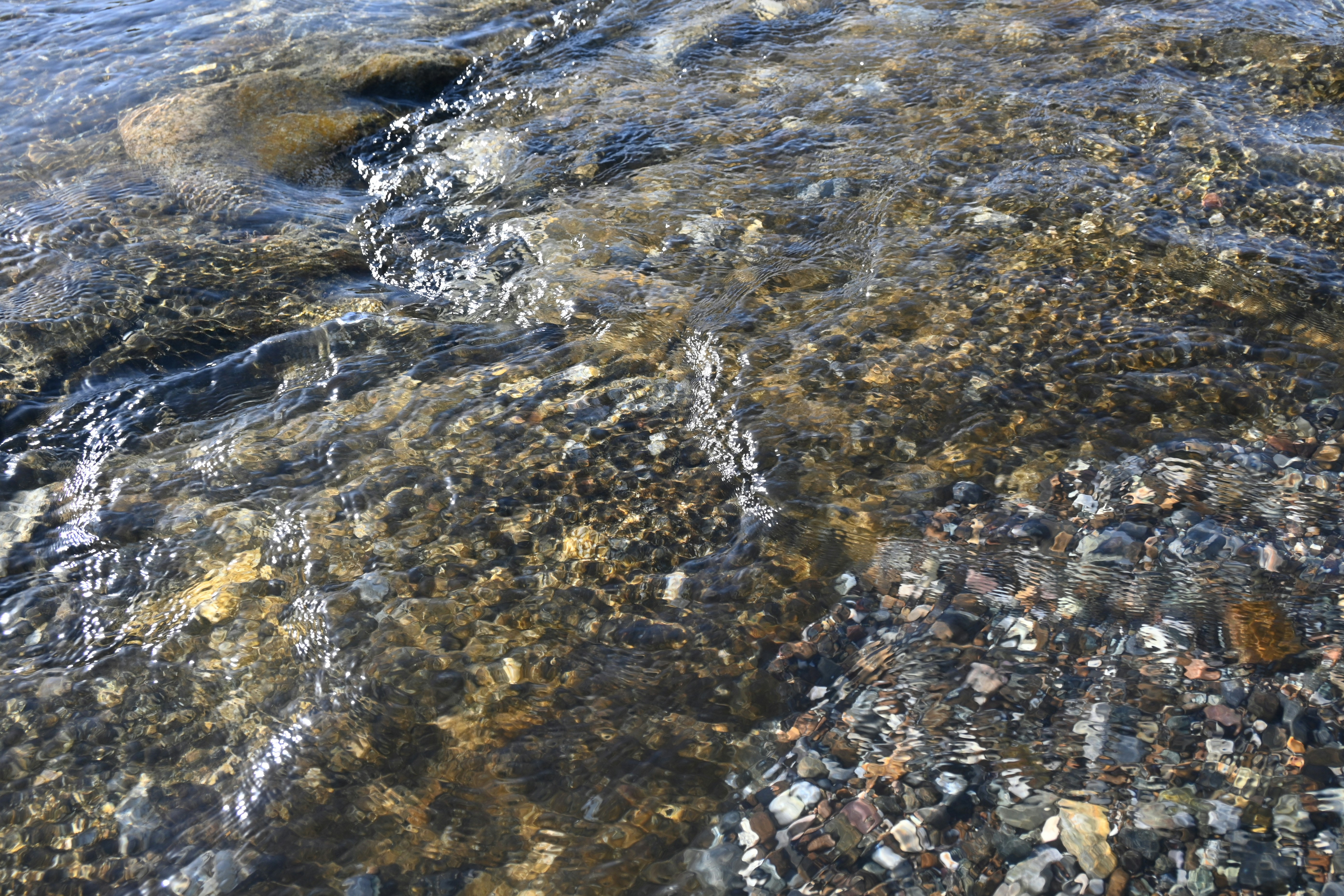 Gros plan sur de l'eau claire et calme qui coule doucement sur des galets ronds et lisses, évoquant la fluidité, le nettoyage et l'apaisement.