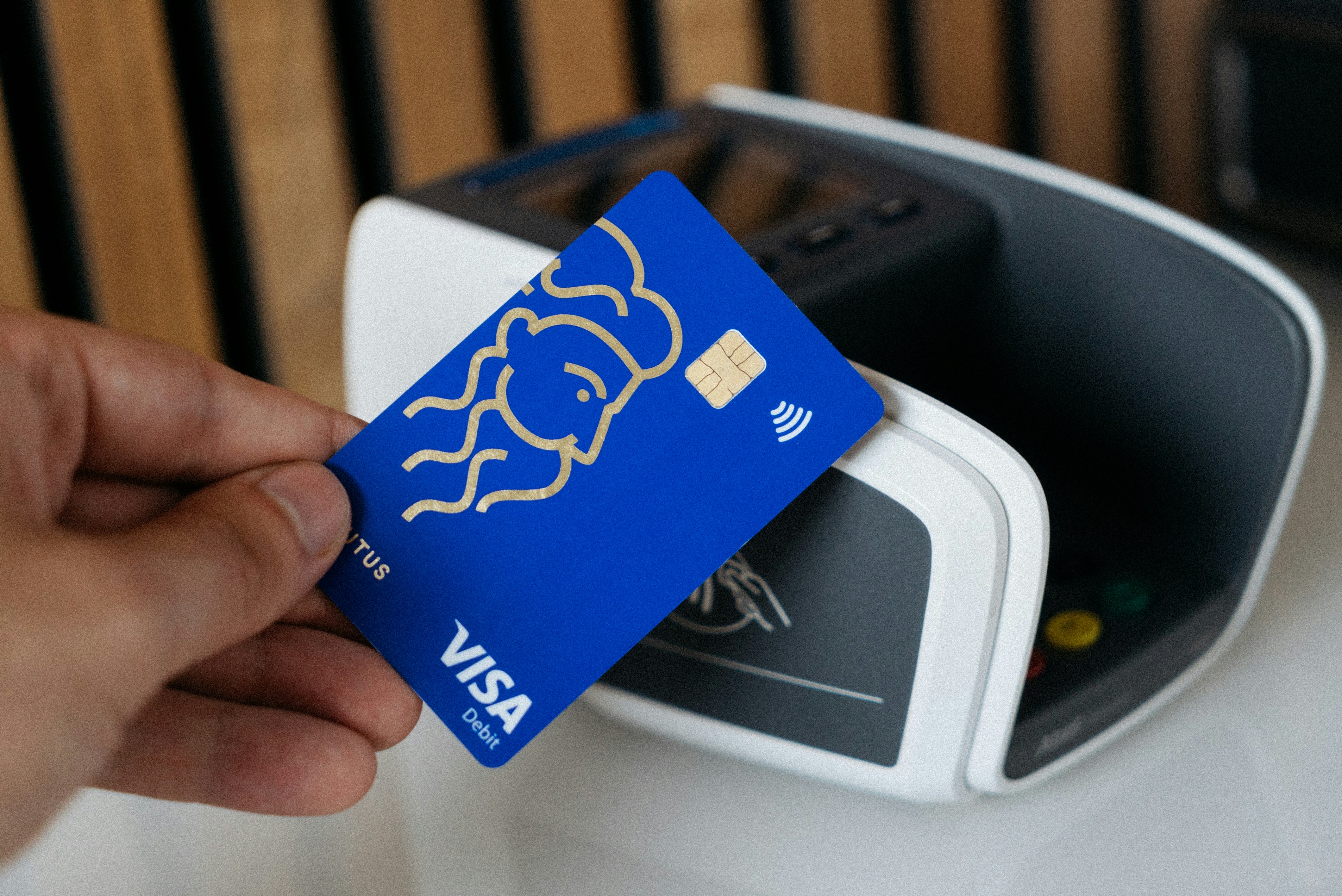 A close-up shot of a hand holding a credit card over a contactless payment terminal, illustrating a 'tap-to-pay' transaction.