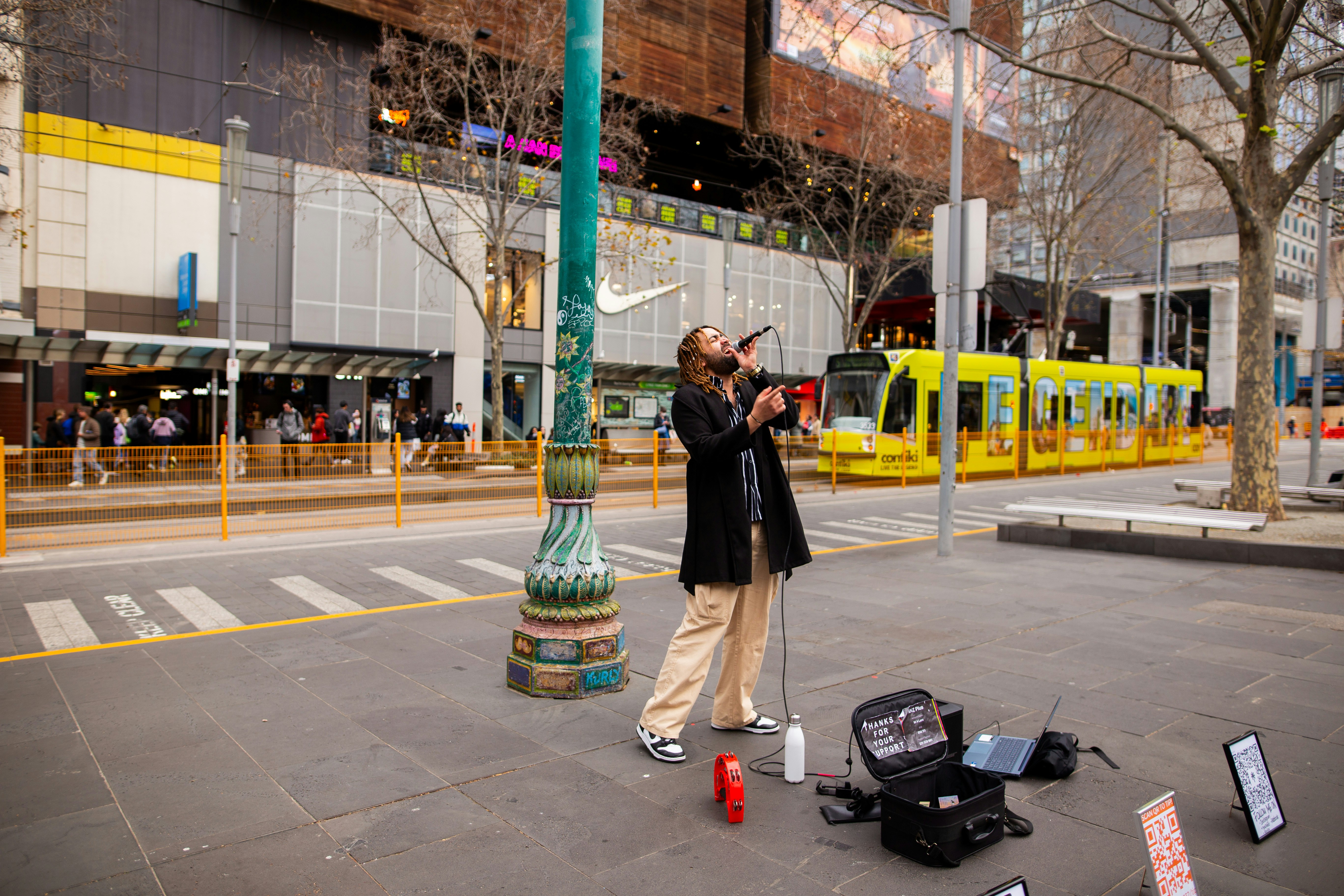 A person's hand holding a smartphone, with the camera app open and focused on a large QR code printed on a sign next to a street performer's guitar case.