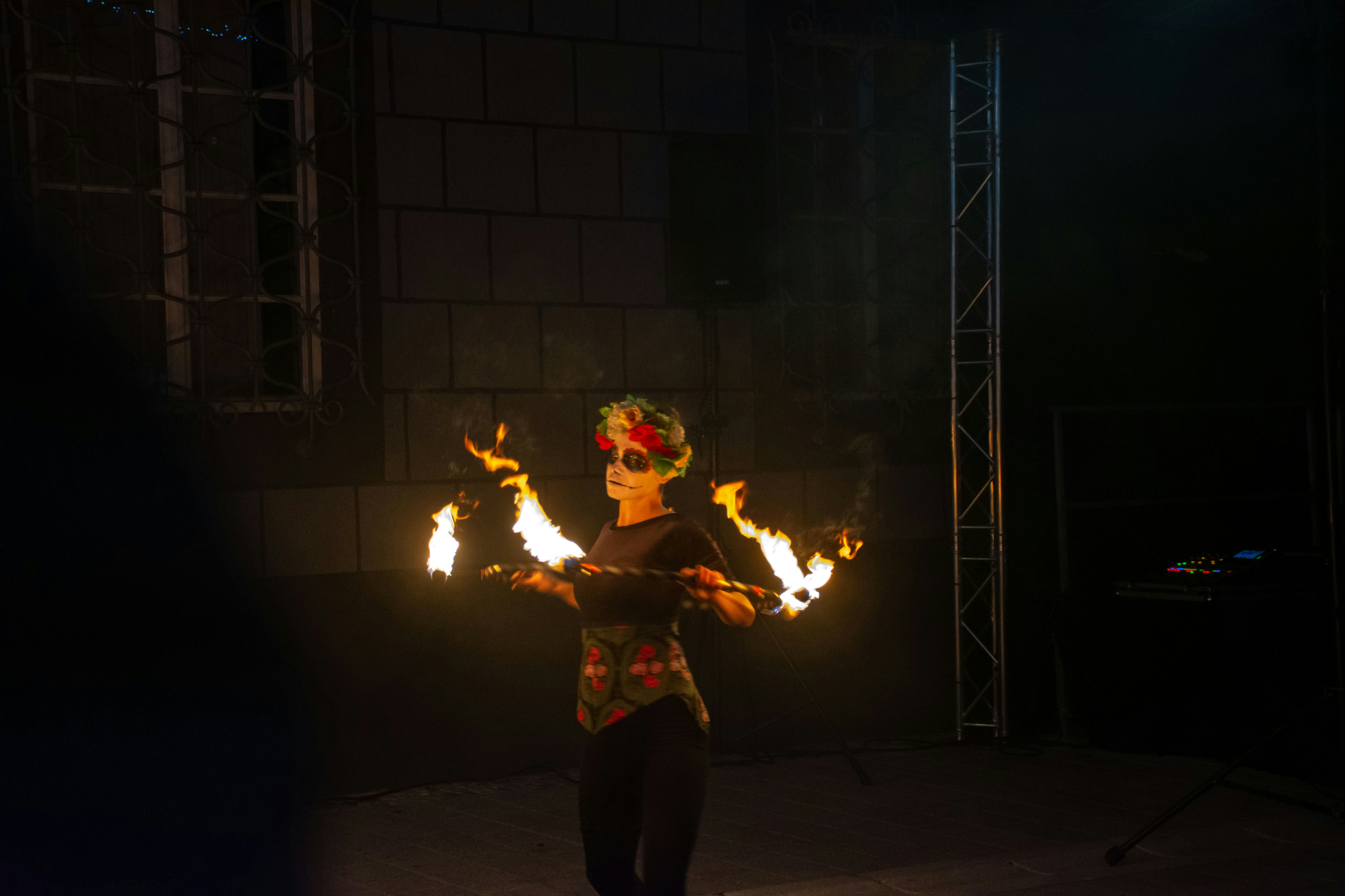 A wide angle image of a performer juggling flaming torches at dusk, with a captivated crowd watching in a park.