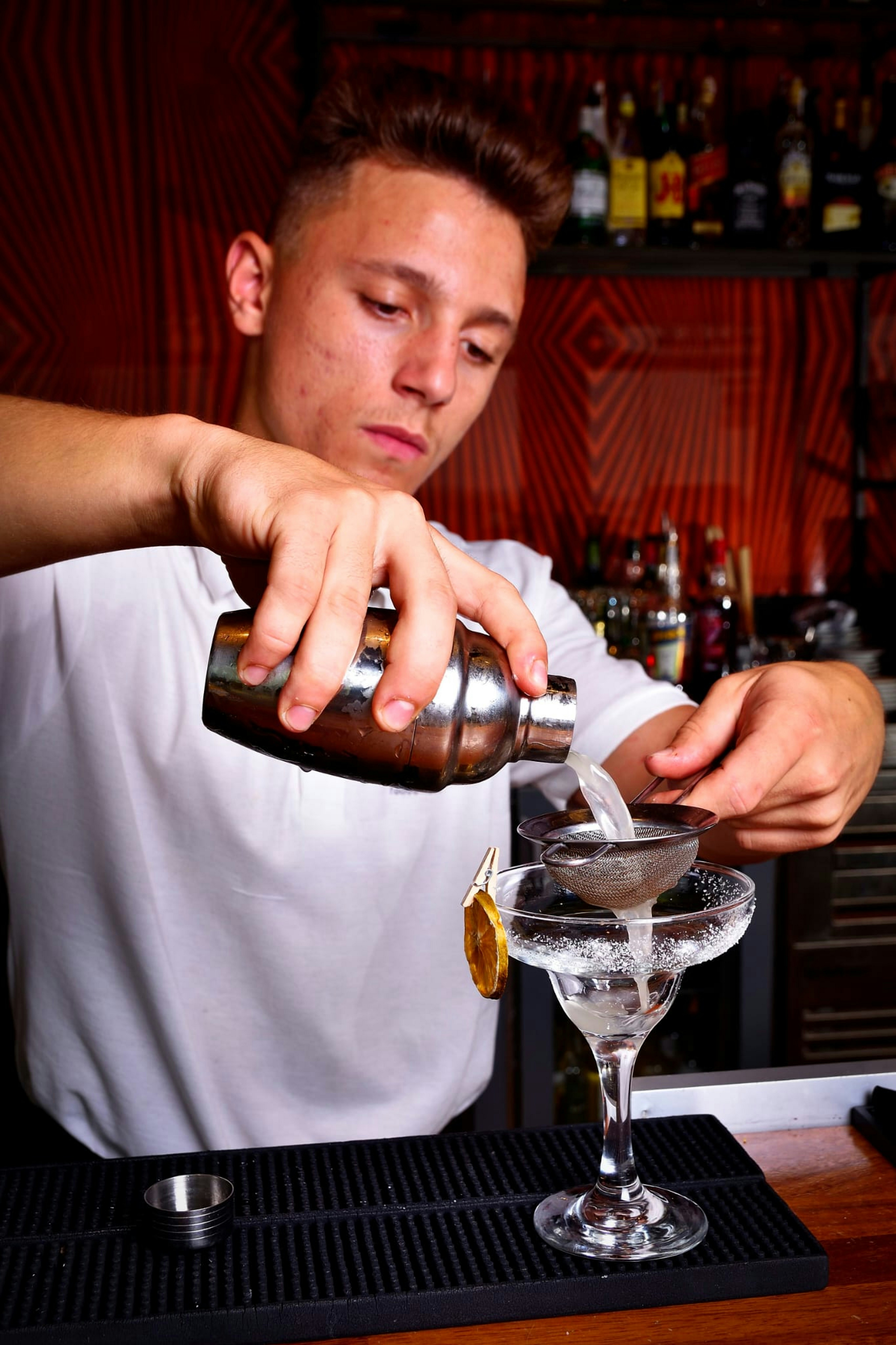 Barman professionnel versant une boisson pétillante dans un verre à cocktail rempli de glace et de garnitures fraîches comme du citron vert et de la menthe.