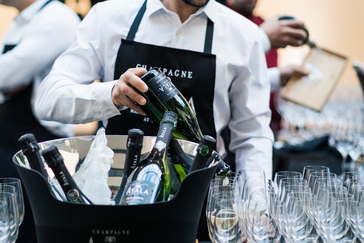 Serveur portant un sac promotionnel de champagne Ayala, préparant des verres pour une dégustation.