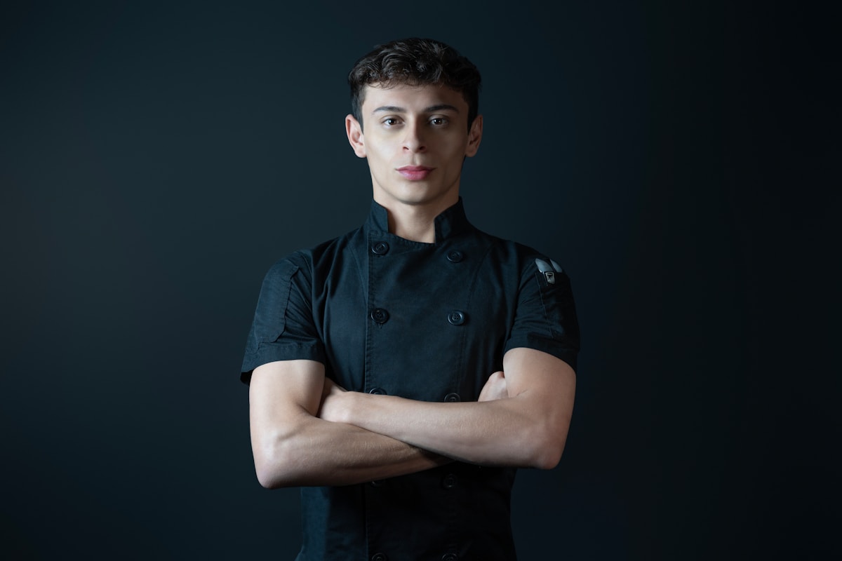 Jeune homme en uniforme de chef, bras croisés, expression sérieuse.