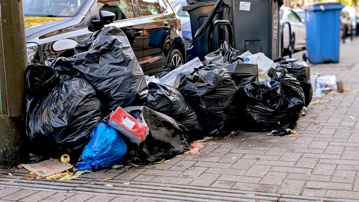 Sacs poubelle noirs empilés sur le trottoir encombré.