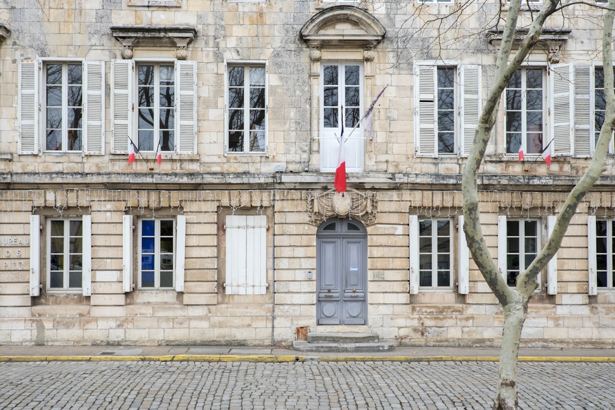 Bâtiment historique en pierre avec fenêtres à volets, drapeaux français.