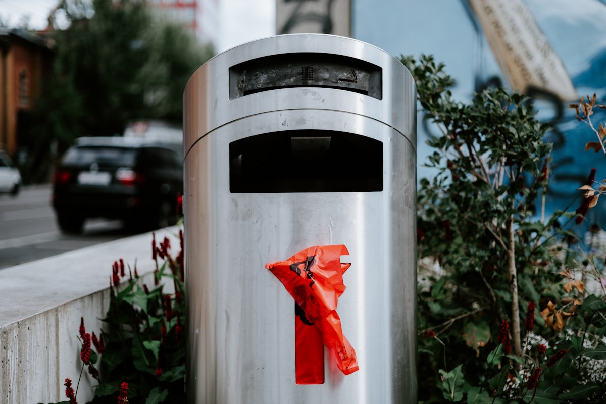 Poubelle métallique avec un sac rouge froissé à l'intérieur, entourée de plantes.
