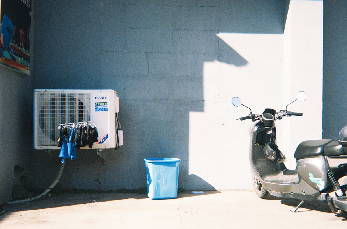 Mur gris, climatiseur, scooter, poubelle bleue, ombre lumineuse.