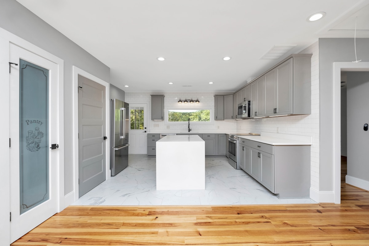 Cuisine moderne avec armoires grises, îlot central blanc et plancher en bois.