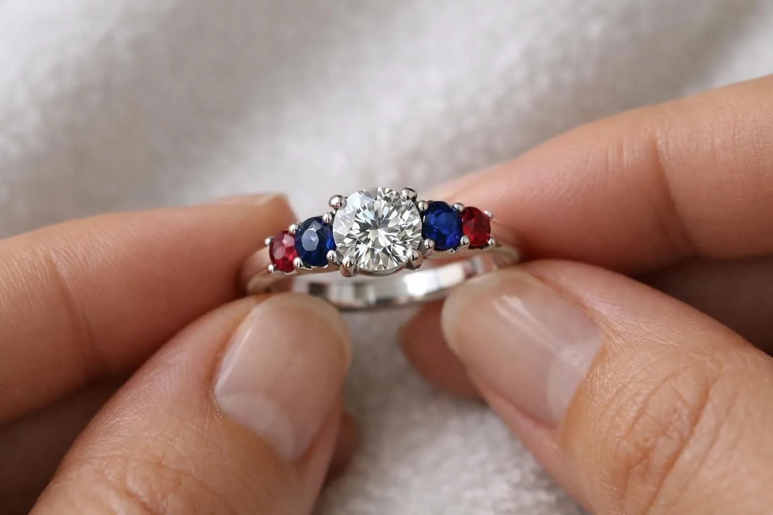 Bague de fiançailles en or blanc avec diamant taille brillant de 2 carats entouré de quatre saphirs bleus et deux rubis rouges montés en pavé, photographie macro professionnelle sur fond de velours blanc, éclairage joaillier latéral révélant les facettes et la profondeur des gemmes