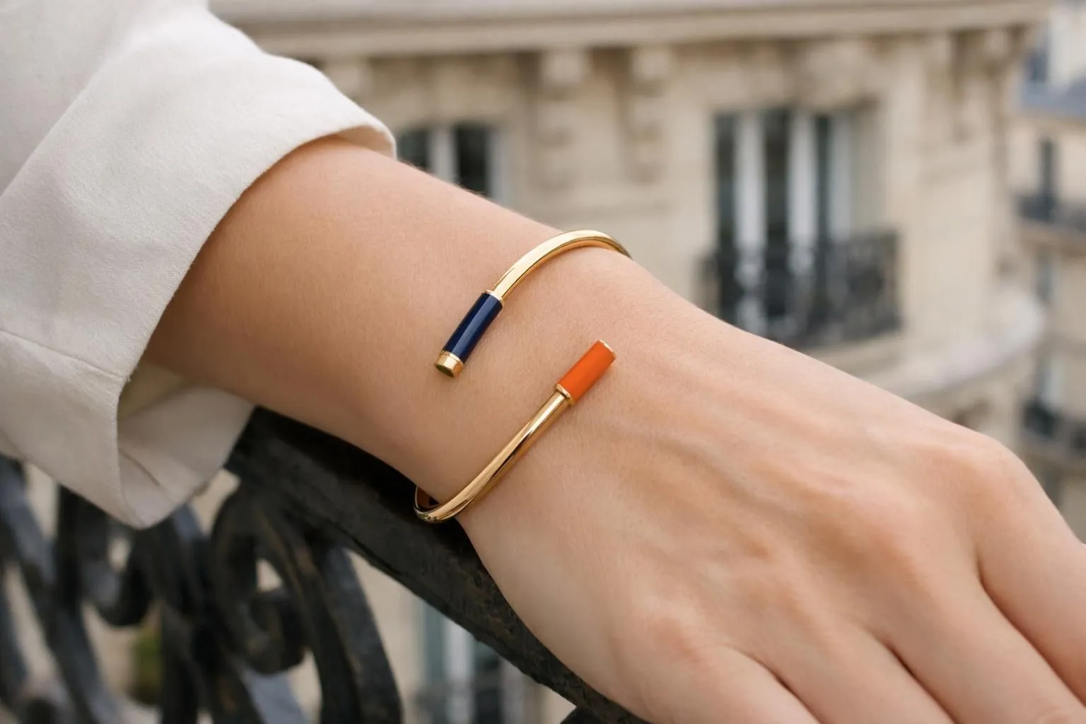 Close-up of an elegant Parisian woman's wrist wearing a luxurious solid gold bangle bracelet against a backdrop of Haussmannian architecture balcony with soft natural lighting highlighting the jewelry's refined craftsmanship