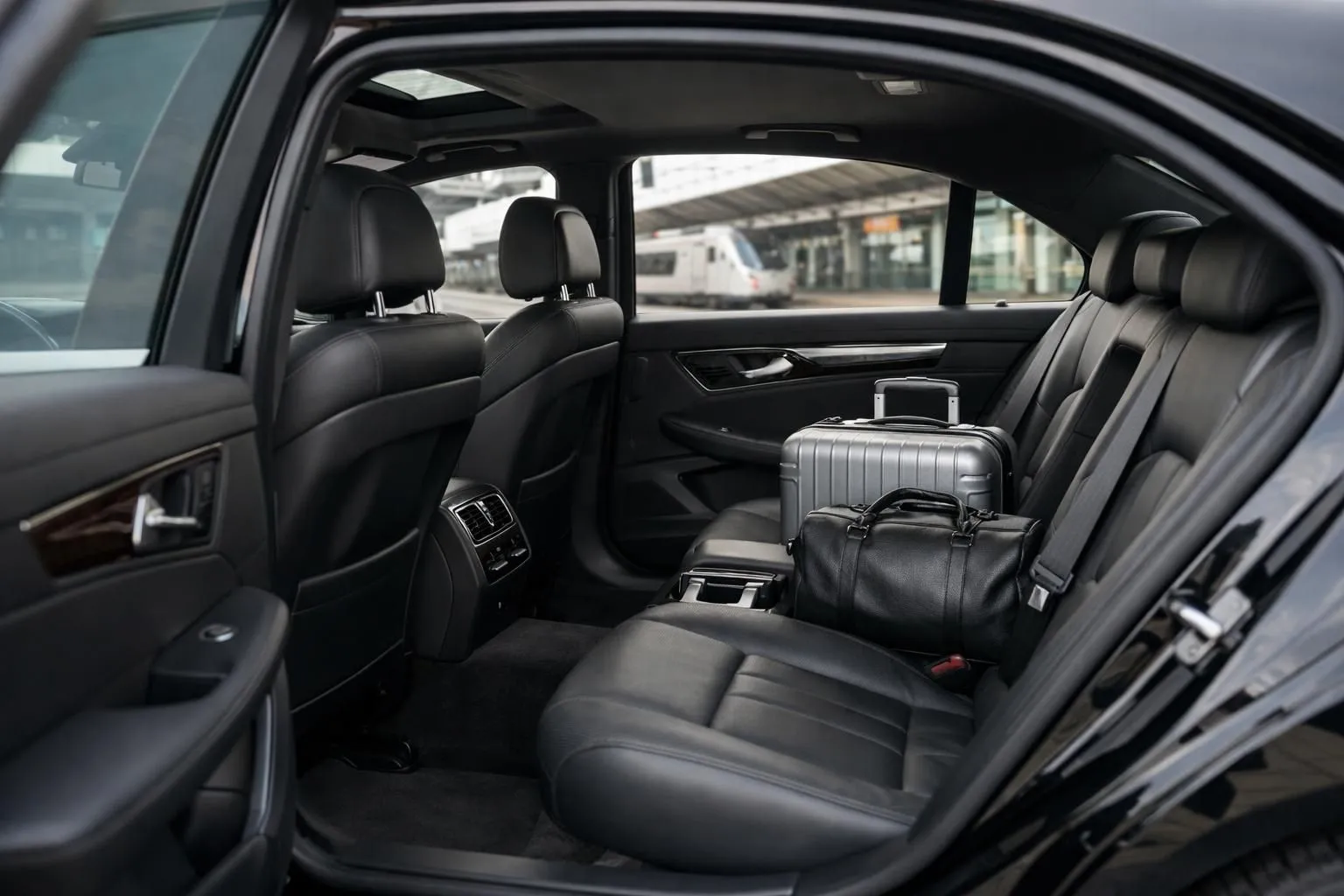 Elegant black sedan vehicle interior with premium leather seats, parked near a modern train station platform, showcasing professional chauffeur service with luggage compartment visible, natural daylight through windows, realistic photography style for Nancy to Paris transportation service