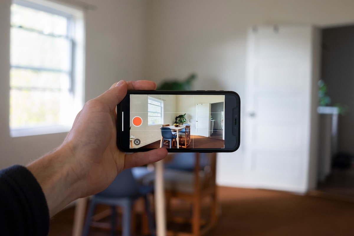 Une main tenant un smartphone capturant l'intérieur d'une pièce avec une table et des chaises.