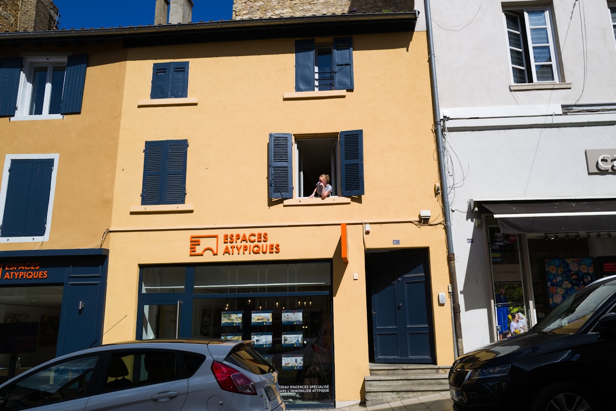 Bâtiment jaune avec fenêtres bleues, enseigne "Espaces atypiques", voitures garées devant.