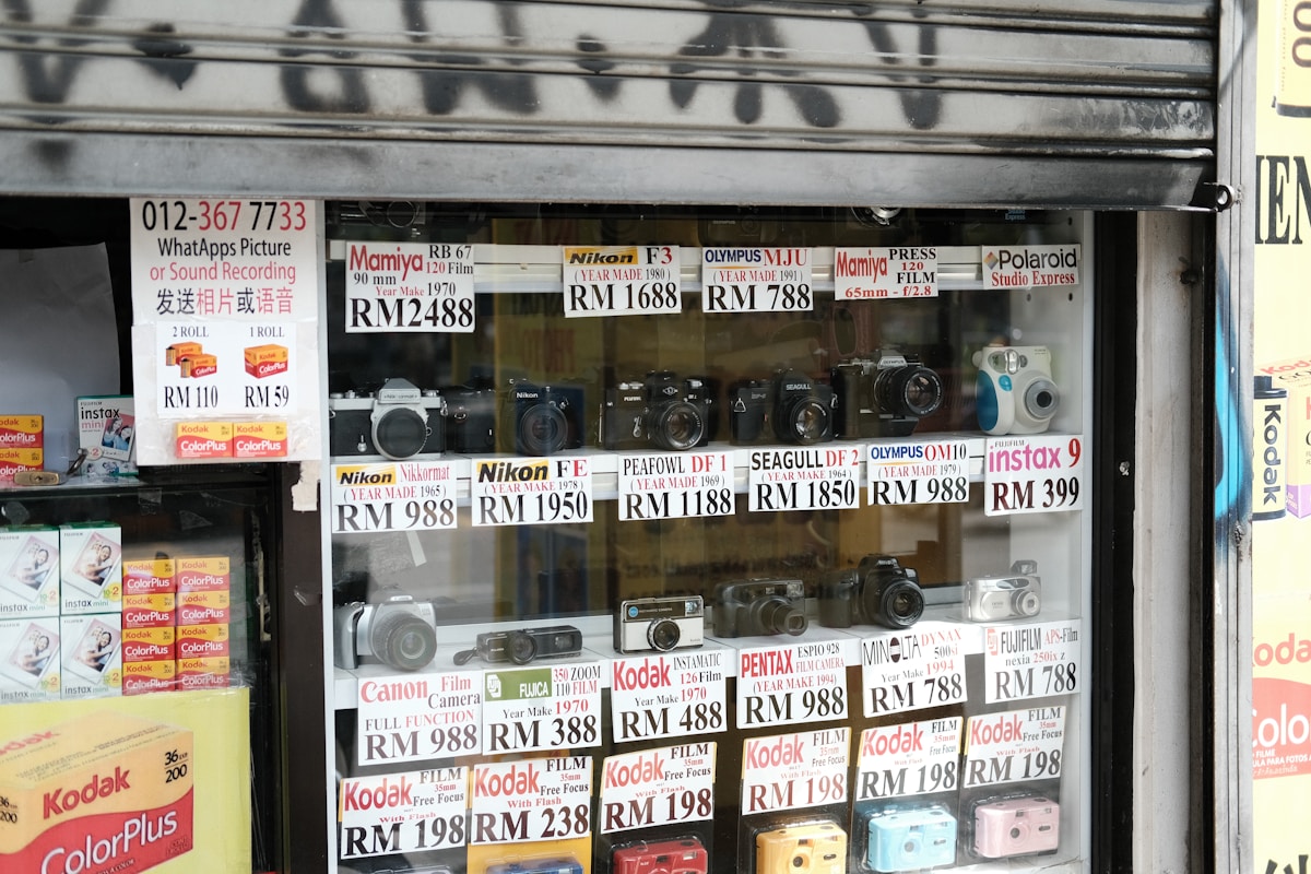 Vitrine de magasin d'appareils photo et accessoires, avec différents modèles et prix affichés.