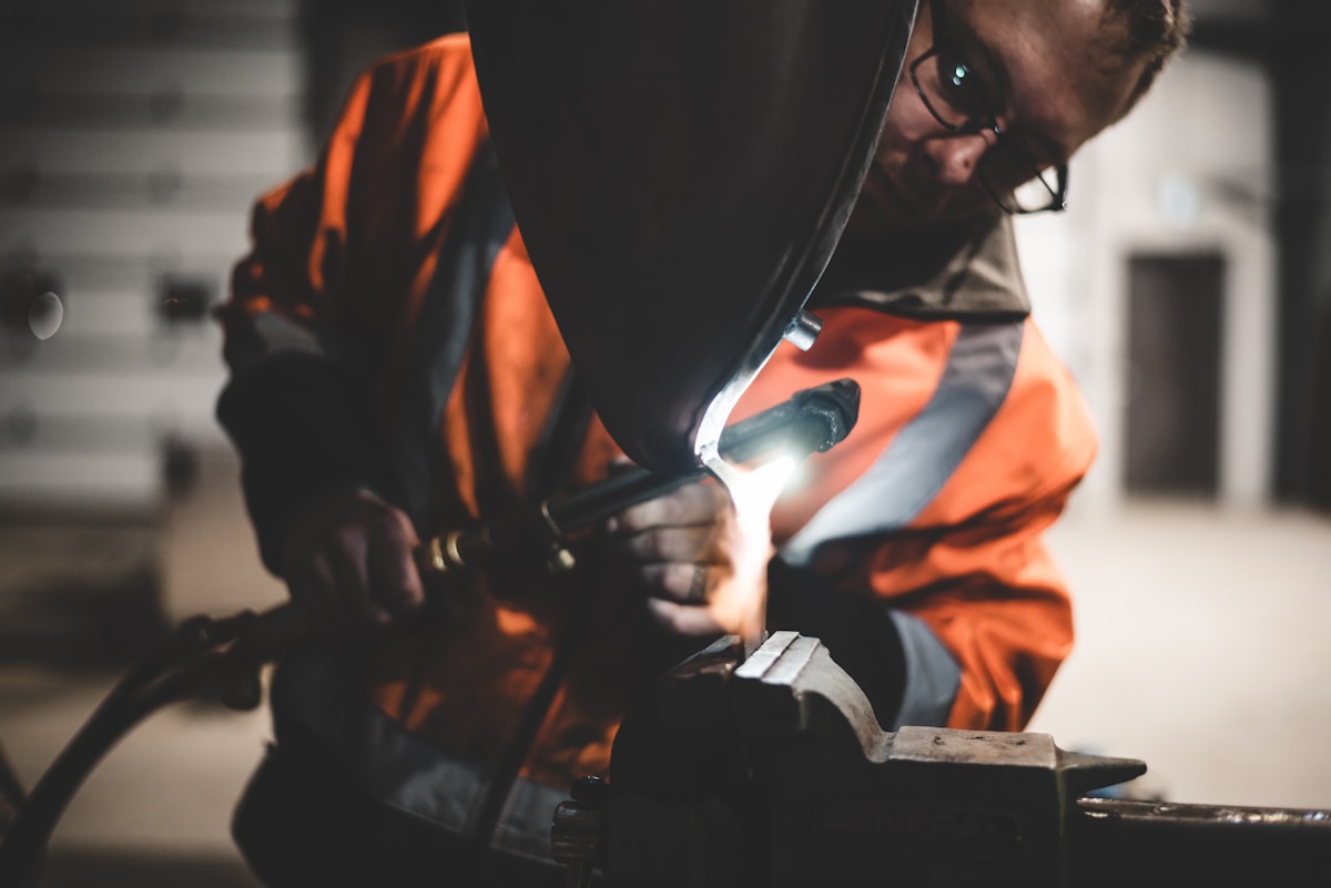 Travailleur en tenue de sécurité orange soudant un métal dans un atelier sombre.