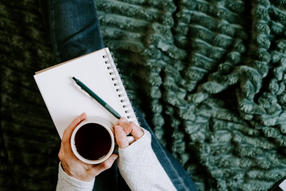 Une personne tenant une tasse de café à côté d'un cahier et d'un stylo.