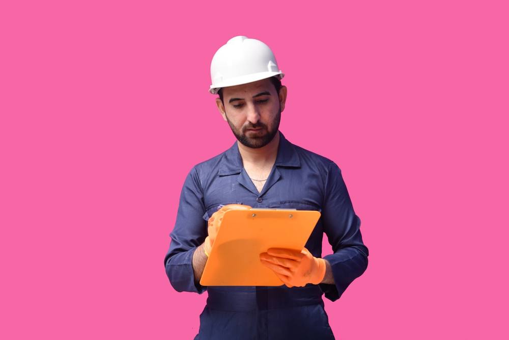 Homme en combinaison bleue et casque blanc tenant un dossier orange.