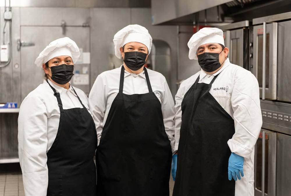 Trois chefs cuisiniers portant des masques et des tabliers noirs dans une cuisine.
