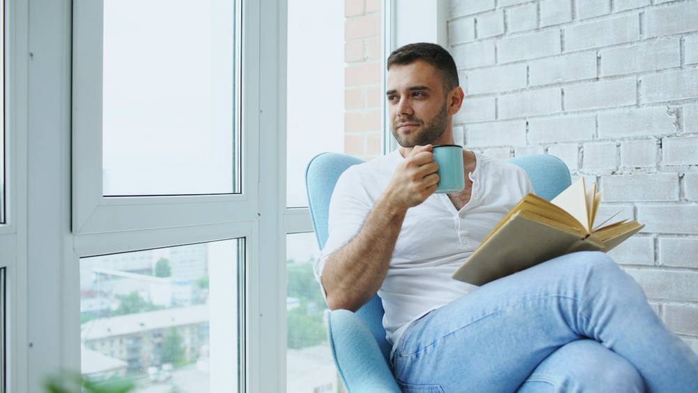 Un homme boit un café et lit un livre près d'une fenêtre.