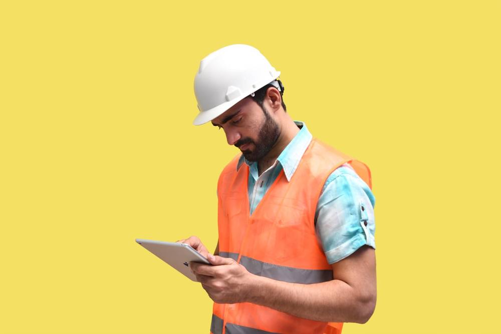 Homme en tenue de sécurité utilisant une tablette numérique sur fond jaune.
