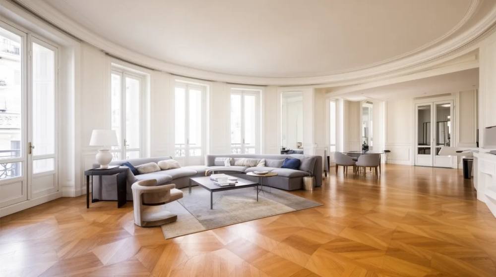 Vaste salon lumineux avec fenêtres panoramiques, mobilier moderne et parquet en bois.