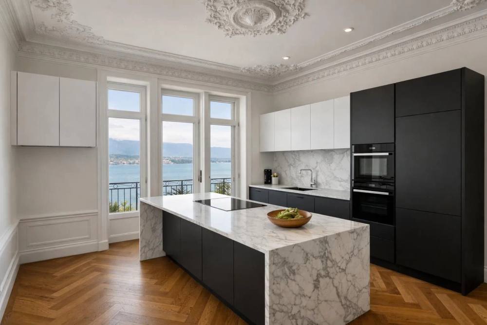 Une cuisine de luxe rénovée dans un appartement genevois de l'époque de la Belle Époque, avec des moulures de plafond ornées, des plans de travail en marbre modernes, des appareils intégrés et une vue sur le lac Léman à travers de hautes fenêtres avec un par