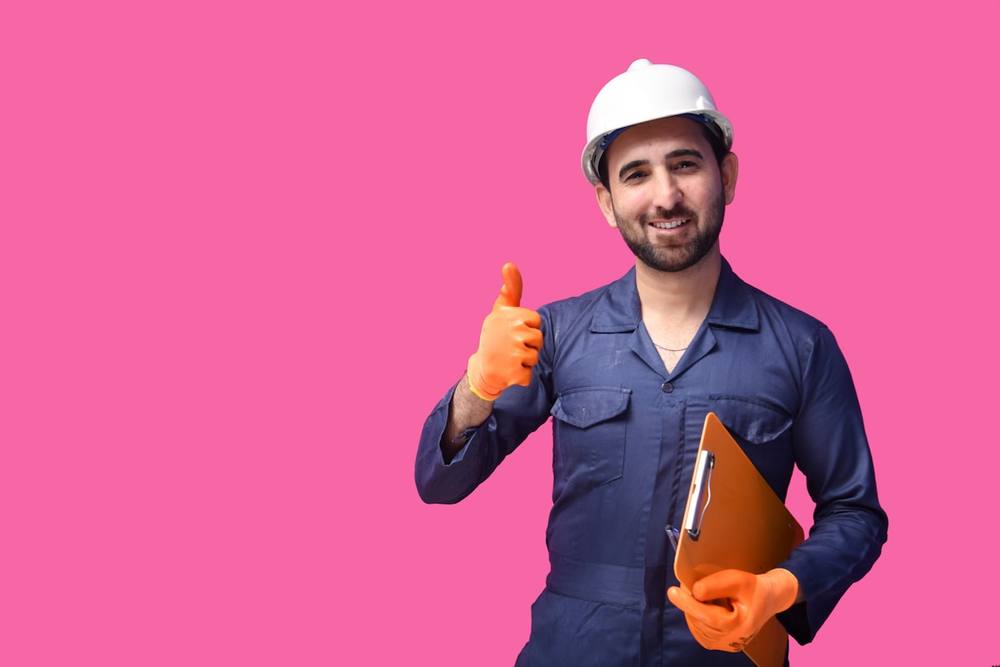 Homme en uniforme de travail donnant un pouce levé sur fond rose.