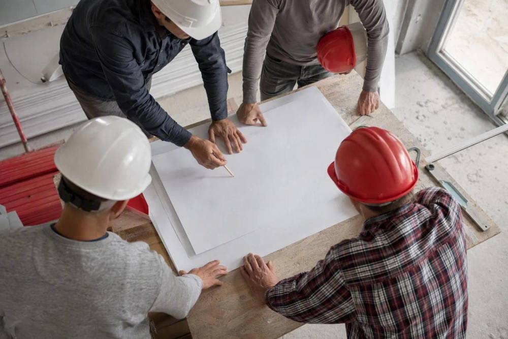 Quatre ouvriers casqués examinent des plans sur une table.