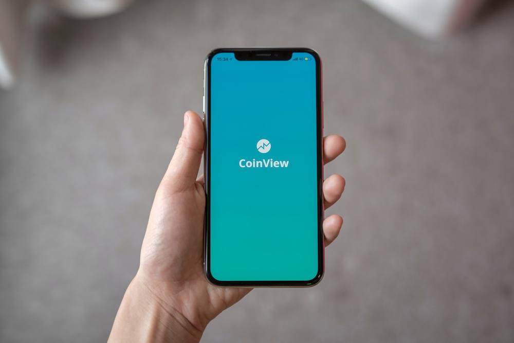 Smartphone affichant l'application CoinView sur un écran bleu turquoise.