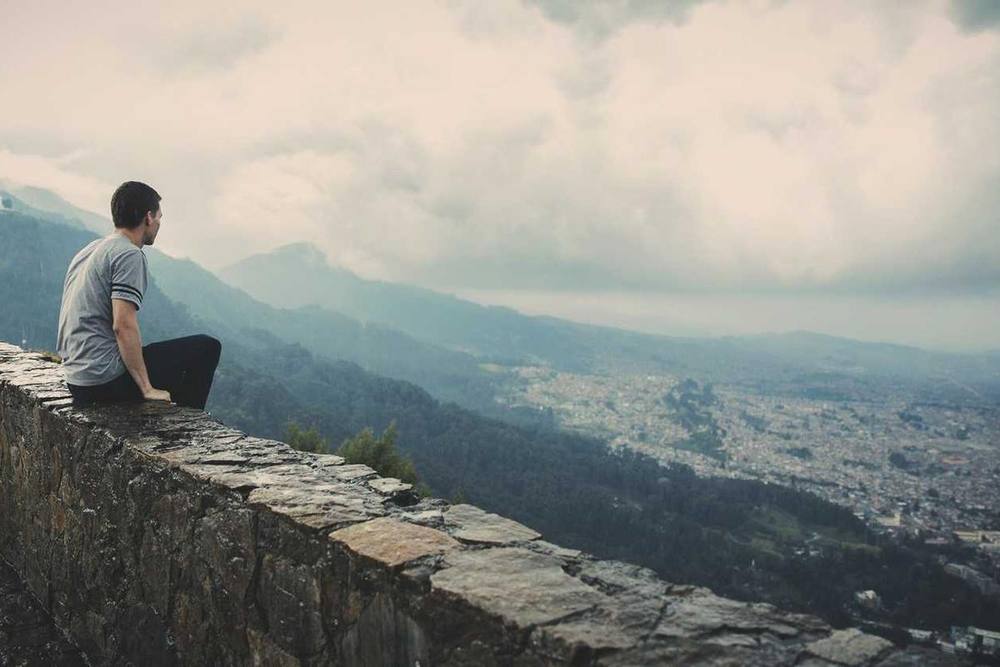 Une personne est assise sur un mur de pierre avec une vue panoramique sur les montagnes et la ville.