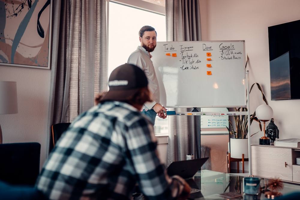 Homme barbu donnant une présentation devant un tableau blanc dans un bureau.